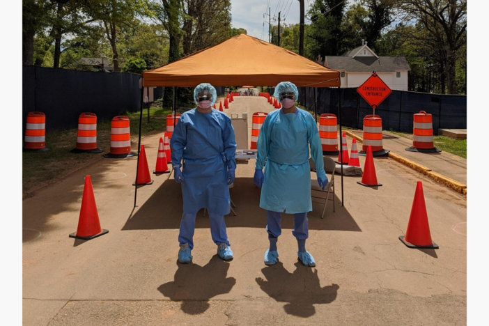 Two doctors with blue hairnets, blue medical scrubs, and masks stand in front of an orange medical tent surrounded by orange traffic cones.