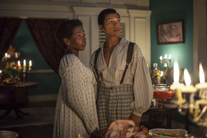A black man and woman in period dress stand in a candlelit dining room.