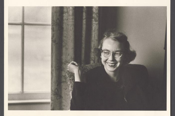 Flannery O’Connor, smiling, sitting by a window.