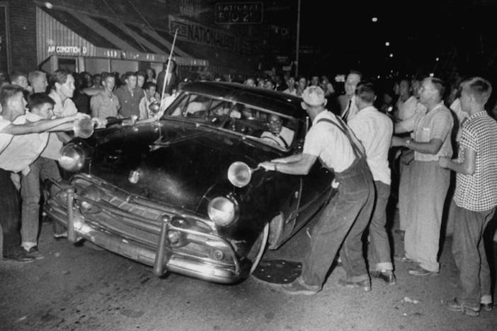 A group of white men attack a car driven by a black man in the 50s.
