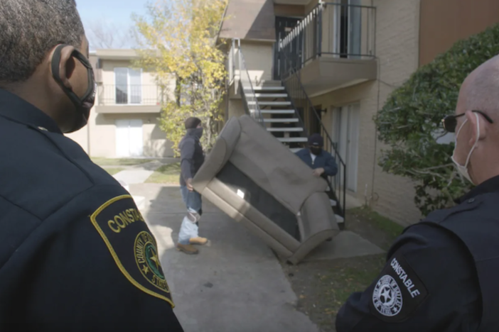 2 police officers watch as men carry a couch down a set of stairs.