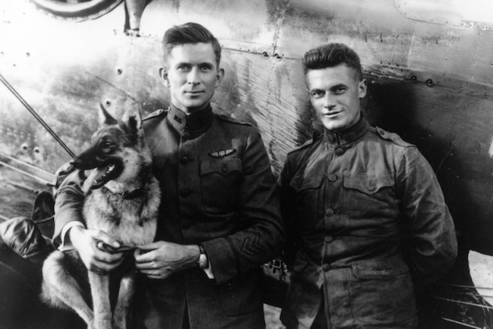 Two men and a dog pose in front of a fighter plane during WWI