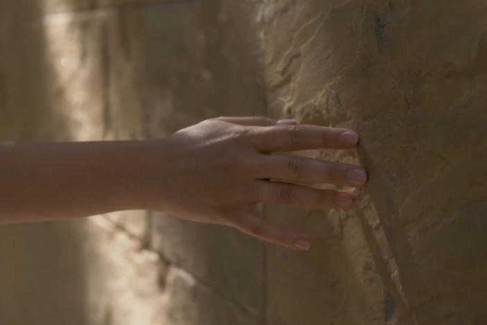 A student hand reaches out to touch a half-lit stone wall in curiosity.
