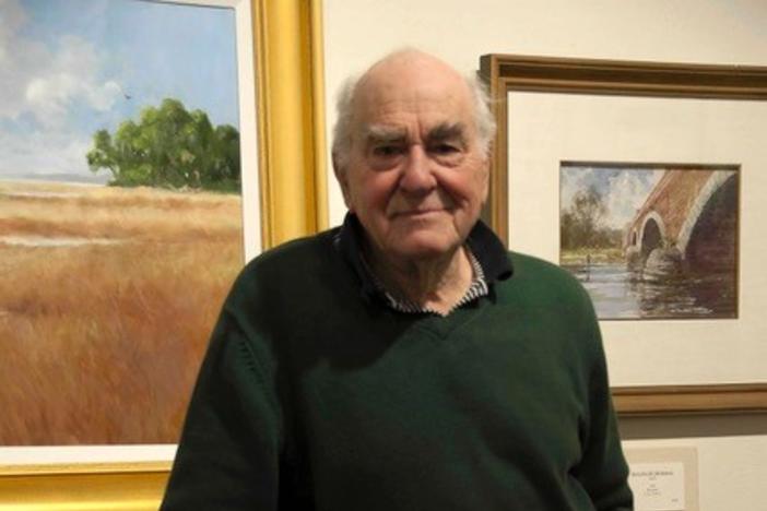 Ray Ellis is shown standing in front of two of his paintings and leaning against a chair in this posed photograph.
