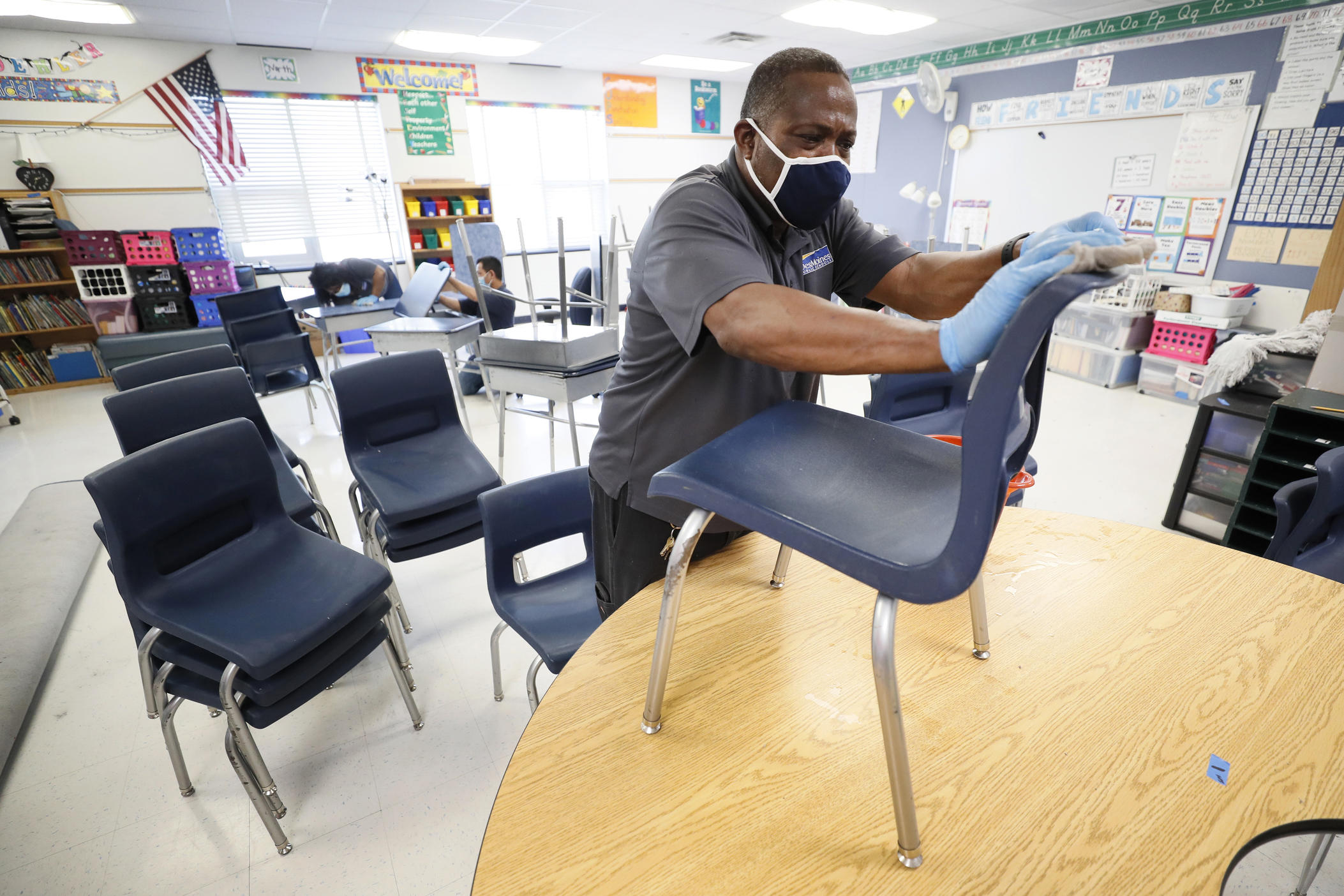 A man in gloves and a mask cleans a chair in a classroom.