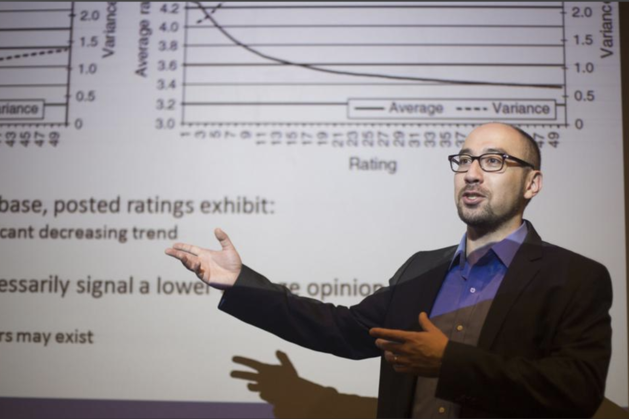 David Schweidel stands in front of a presentation. He's wearing black glasses, a dark suit jacket and a blue button-up shirt.