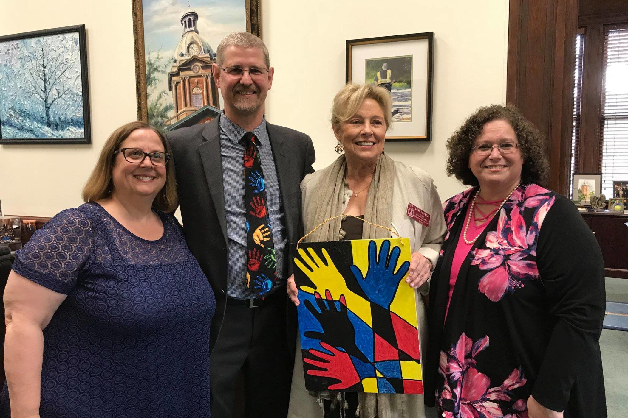 Jimmy Peterson, executive director of the Georgia Center for the Deaf and Hard of Hearing, stands with three women. Everyone is smiling. One woman stands to the left of Jimmy, and she holds a painting of hands in the colors red, yellow and blue.