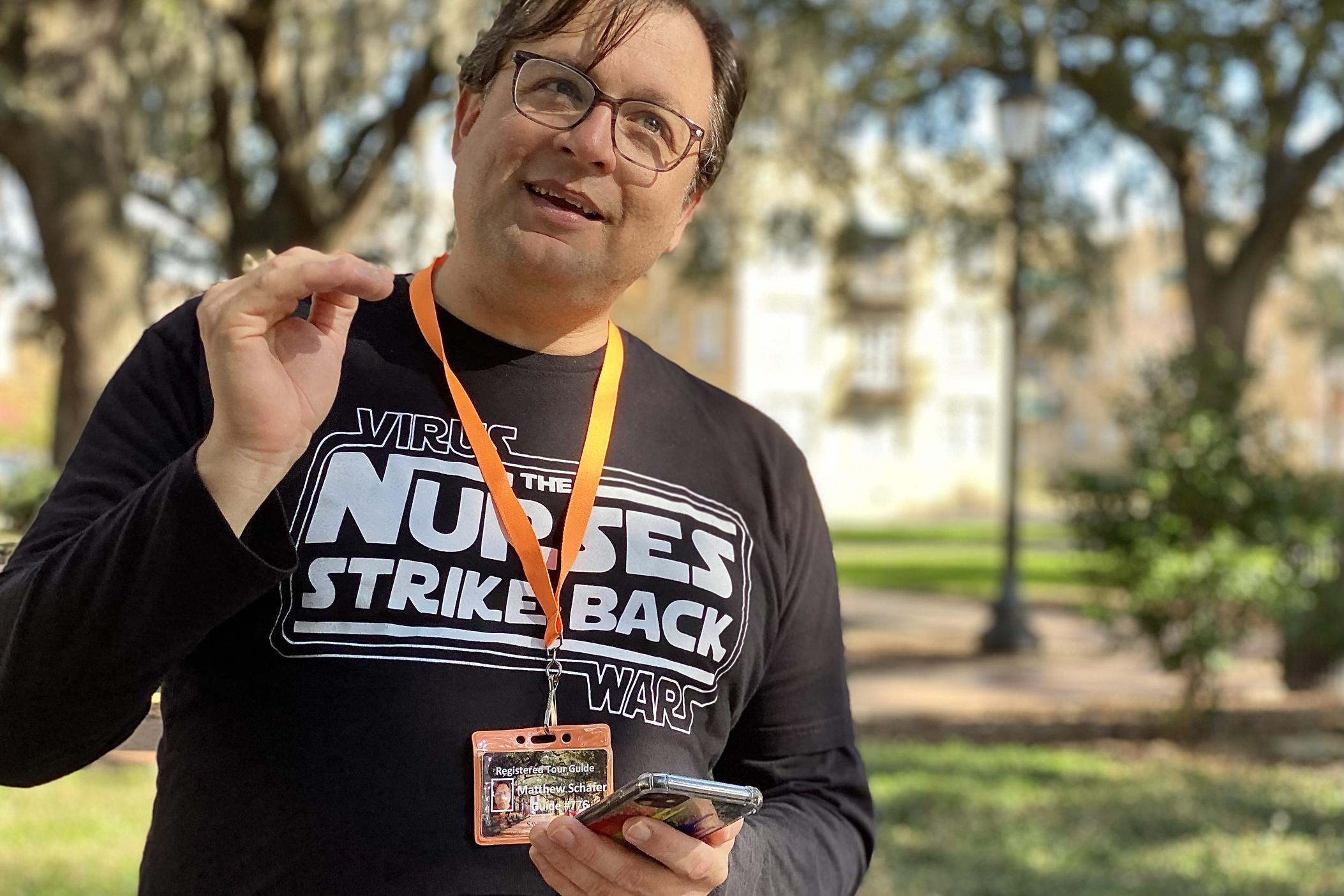 Matt Schafer gestures with his right hand in a park while leading a walking tour about Savannah's medical history. He is wearing a black long-sleeve T-shirt with text that reads “Virus Wars: The Nurses Strike Back.” Around his neck is an orange lanyard that is holding a tour guide identification card.
