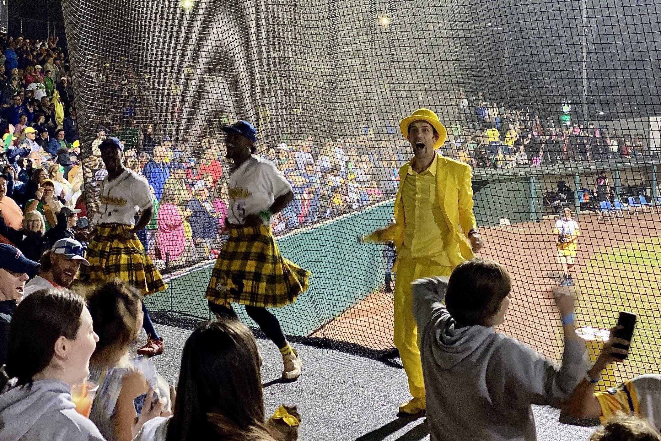 Wearing his signature yellow tuxedo, Savannah Bananas owner Jesse Cole dances with players and fans in between innings at Grayson Stadium. Cole and two players are dancing on top of the home dugout. The players are wearing kilts with a yellow-and-black plaid pattern, in addition to their white home uniforms.
