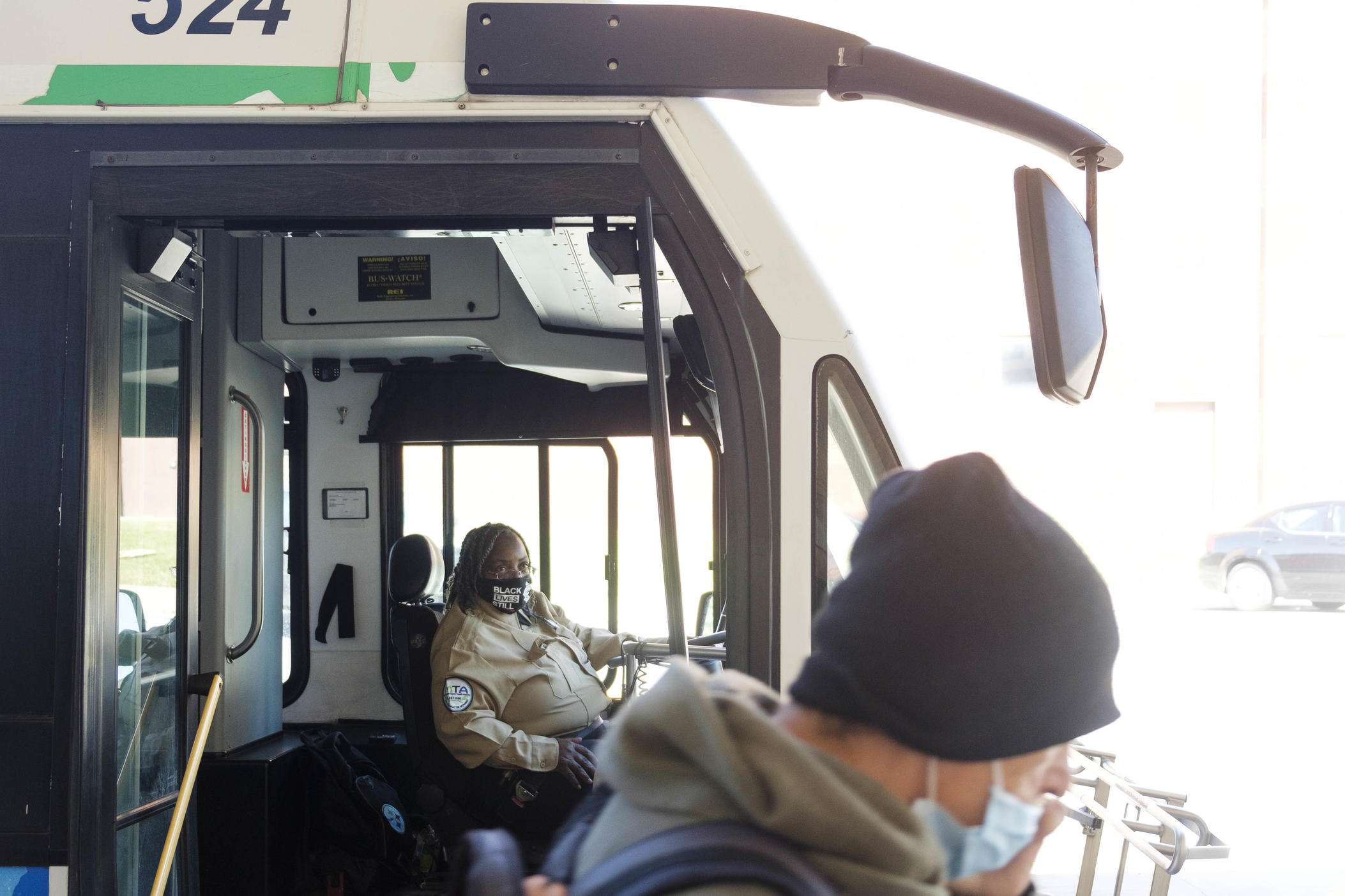 A man wearing a mask walks in front of a bus as the driver opens the door