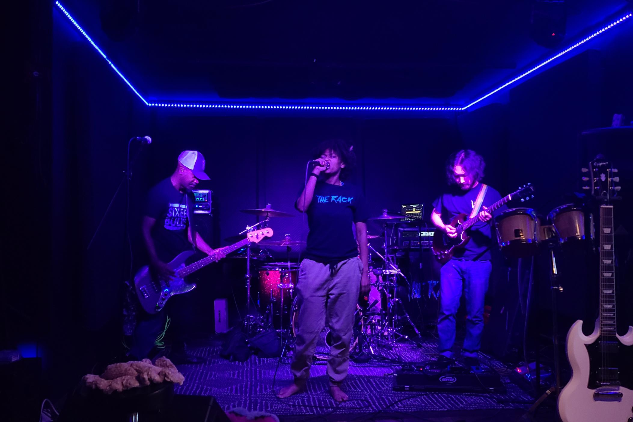 Atlanta-based band The Rack practice for upcoming tour and performance at Afropunk on July 26, 2023. The lead singer is wearinga black shirt which says the rack, behind her is a drummer to her left is a bass player and to the right a guitarist. They're all bathed in purple light.