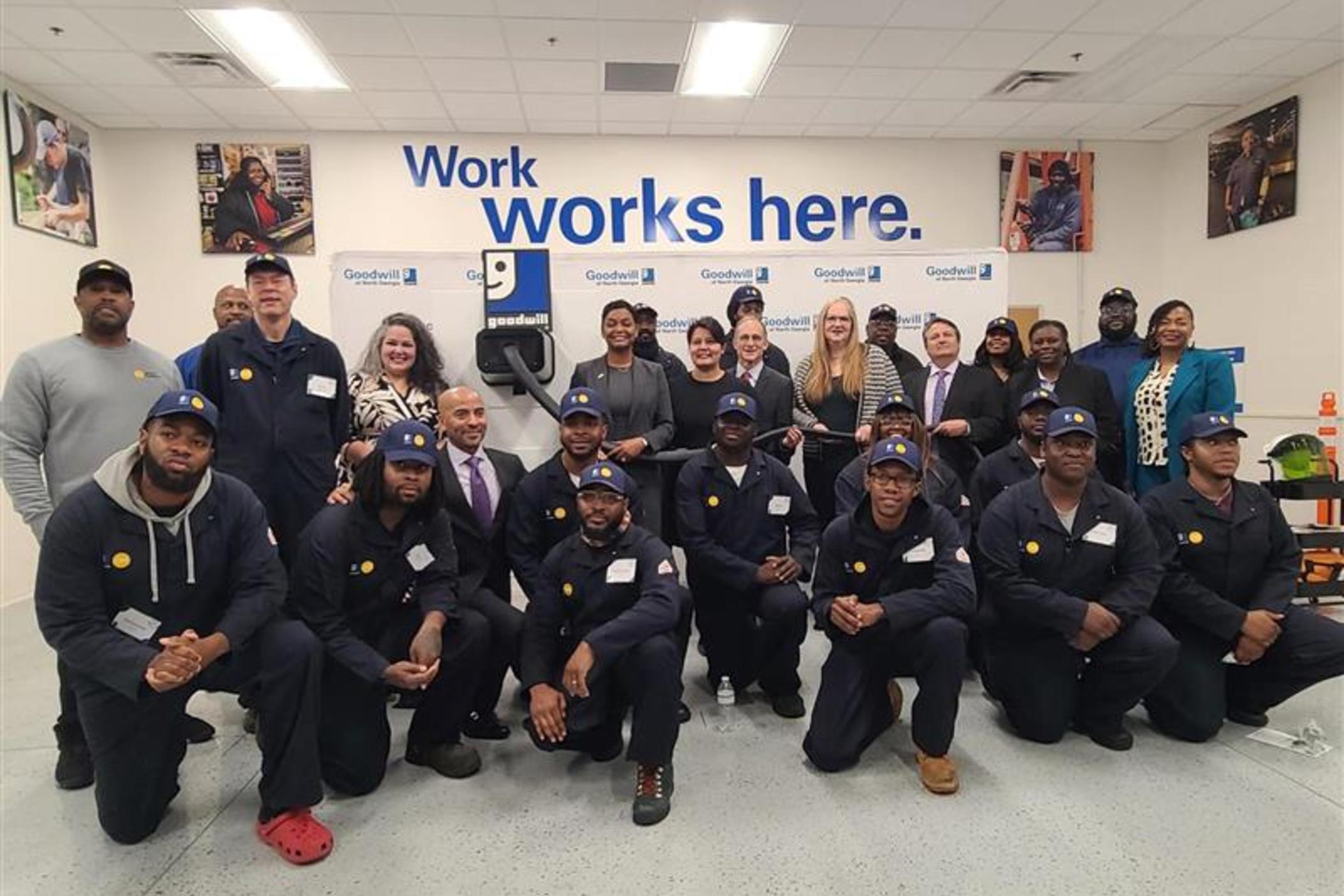 Students of the first Clean Tech Infrastructure Academy cohort gather at the Goodwill to celebrate completing the program January 23, 2024. They each have on blue work jumpsuits and blue hats.