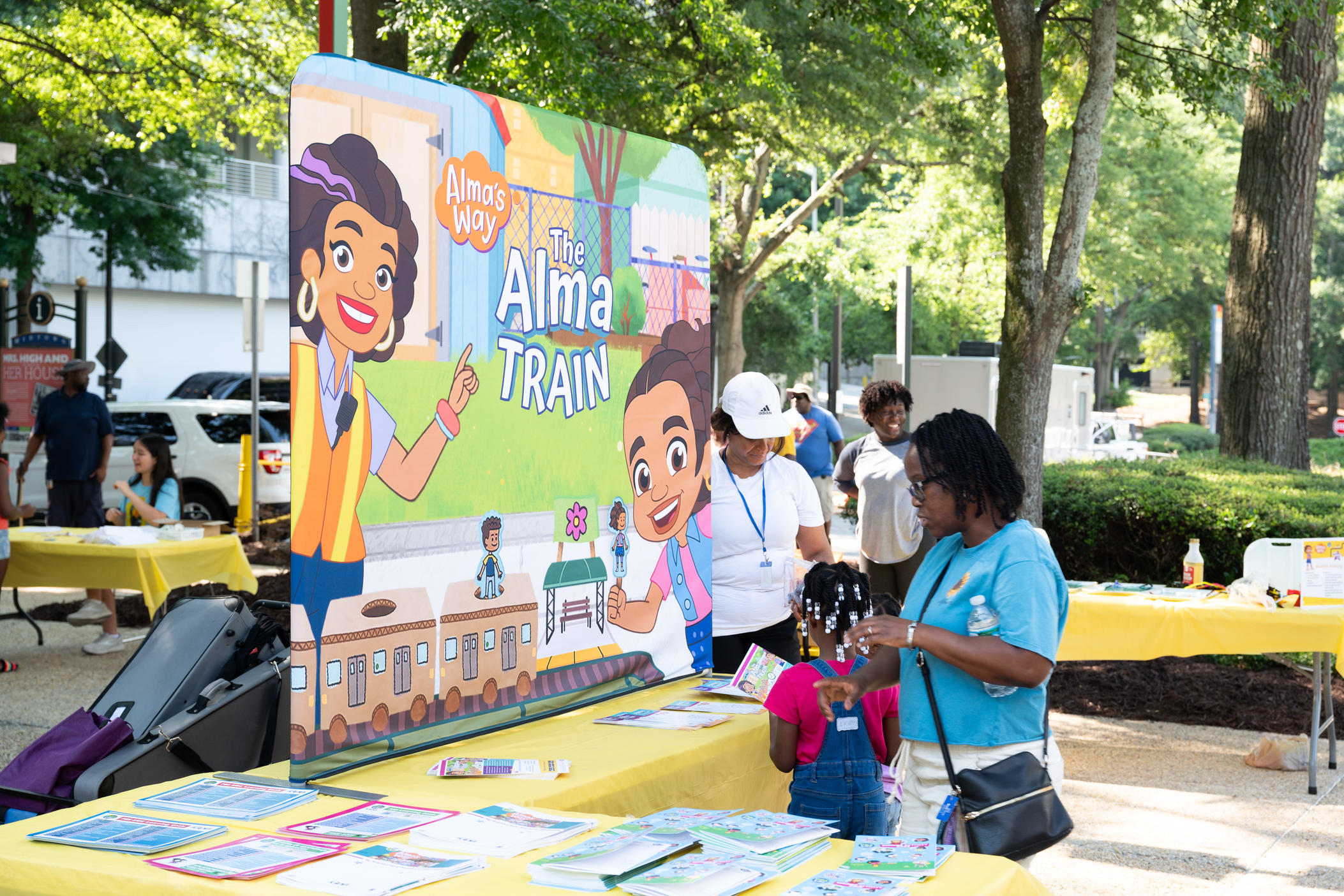 a mother and child stand next to a large poster that says the Alma Train with two cartoon characters smiling and a cartoon train at the bottom of the poster.