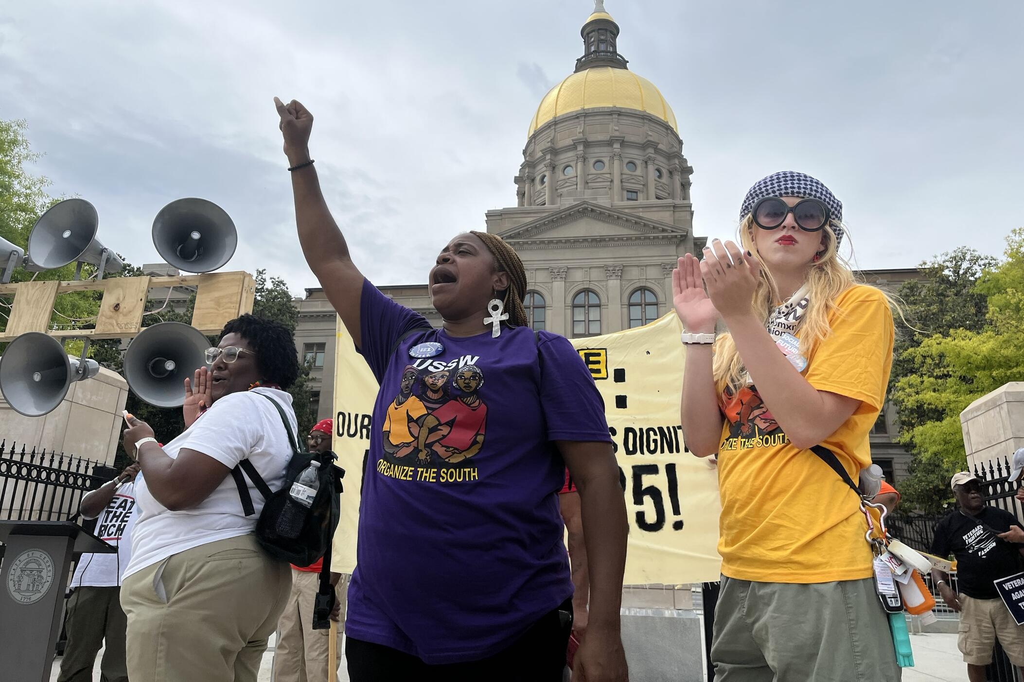 Three women in Union shirts stand in front of the state capitol, one has her first up.