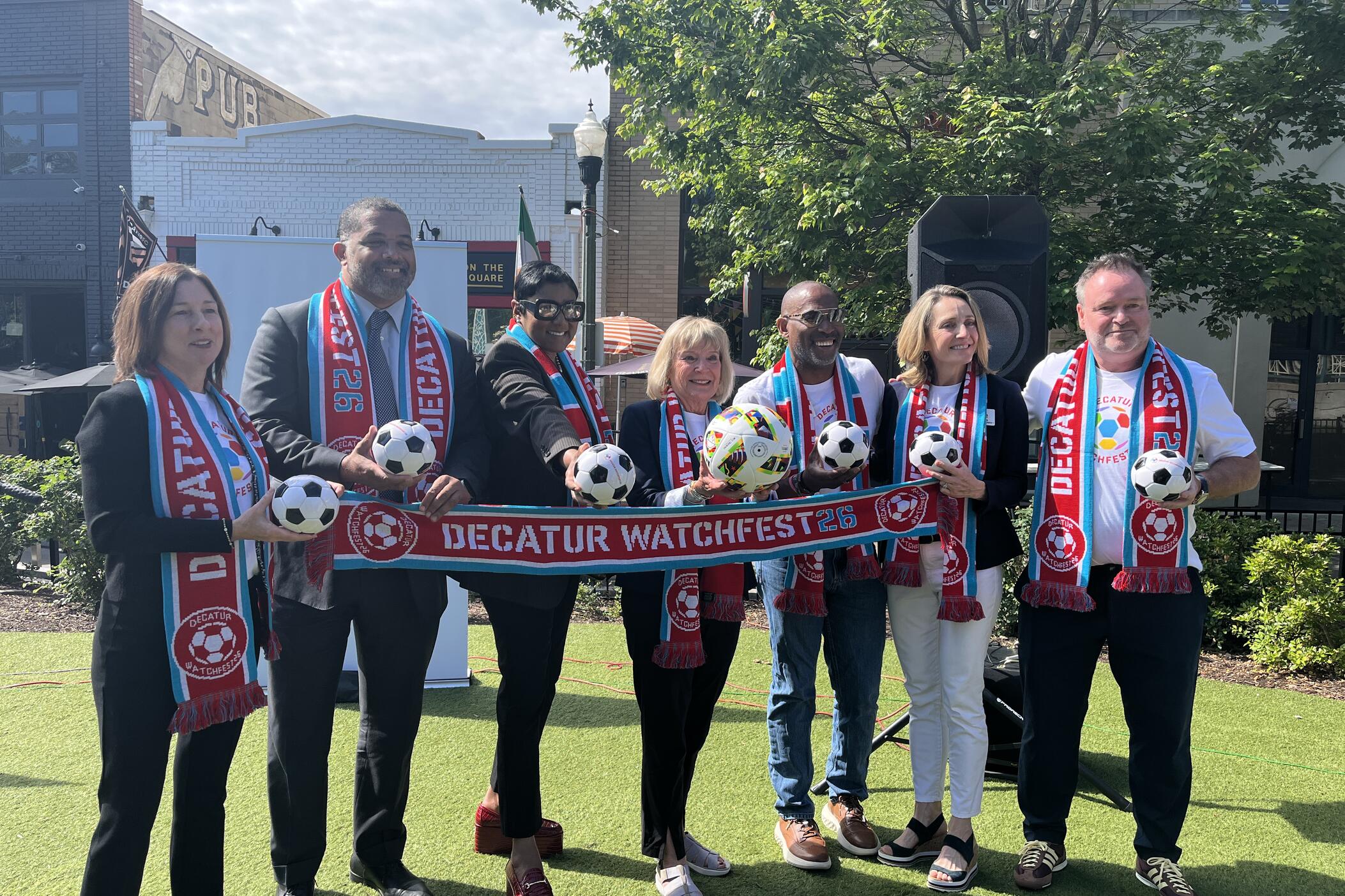 7 people standing behind a scarf that says " Decatur Watchfest" and they're all holding soccer balls and wearing matching scafs
