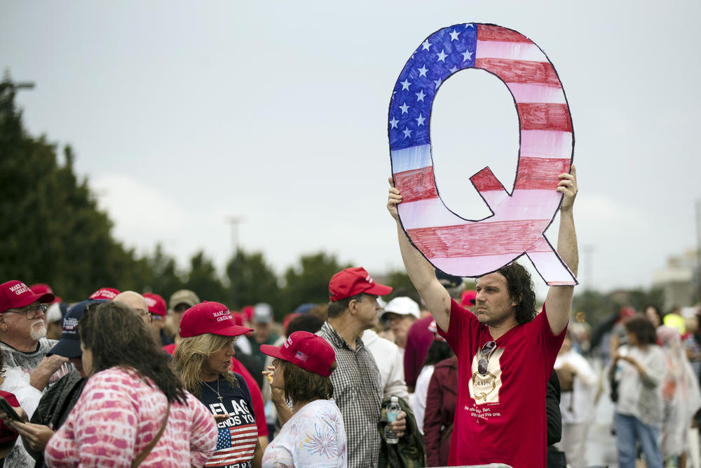 A male protester in a red shirt holds up a large Q-shaped sign, which is colored in with the design and colors of the American flag. Other protesters in the background are wearing red MAGA hats.