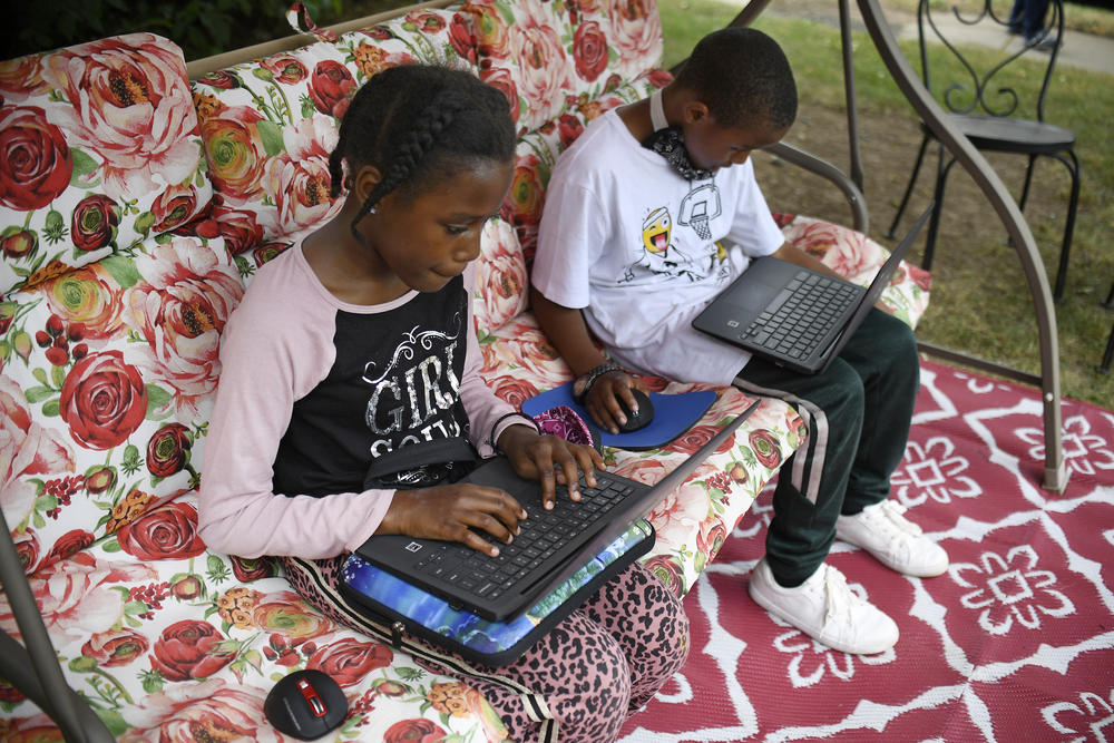 Two young children sit with laptops on a floral couch outside, doing schoolwork.