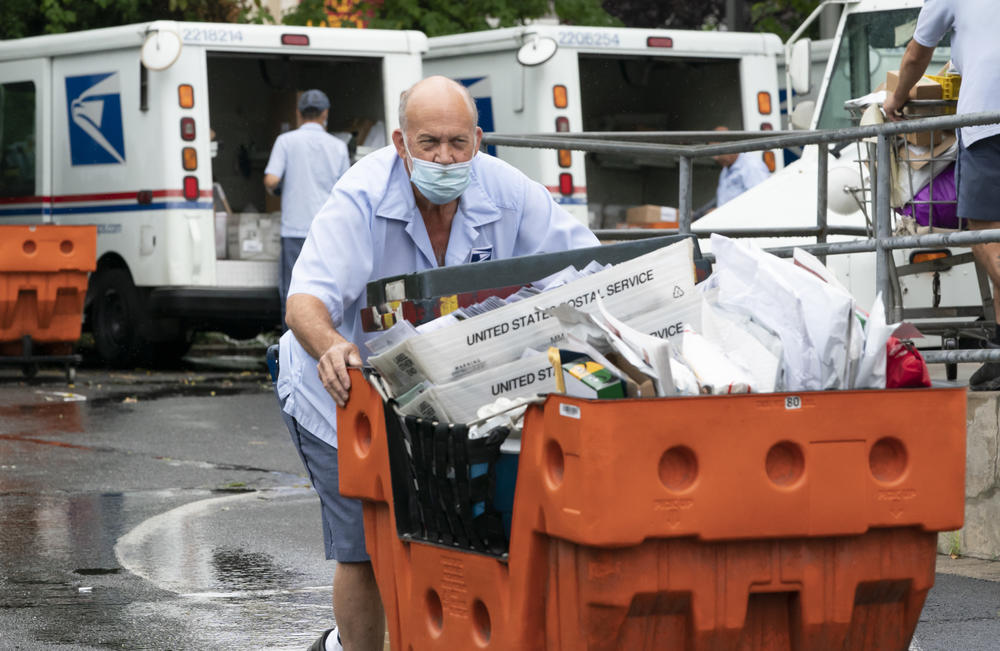 A male U.S. postal worker pushes an orange cart of letter mail towards the camera on a wet day. In the background, two other postal workers unload U.S. Postal Service trucks of mail.