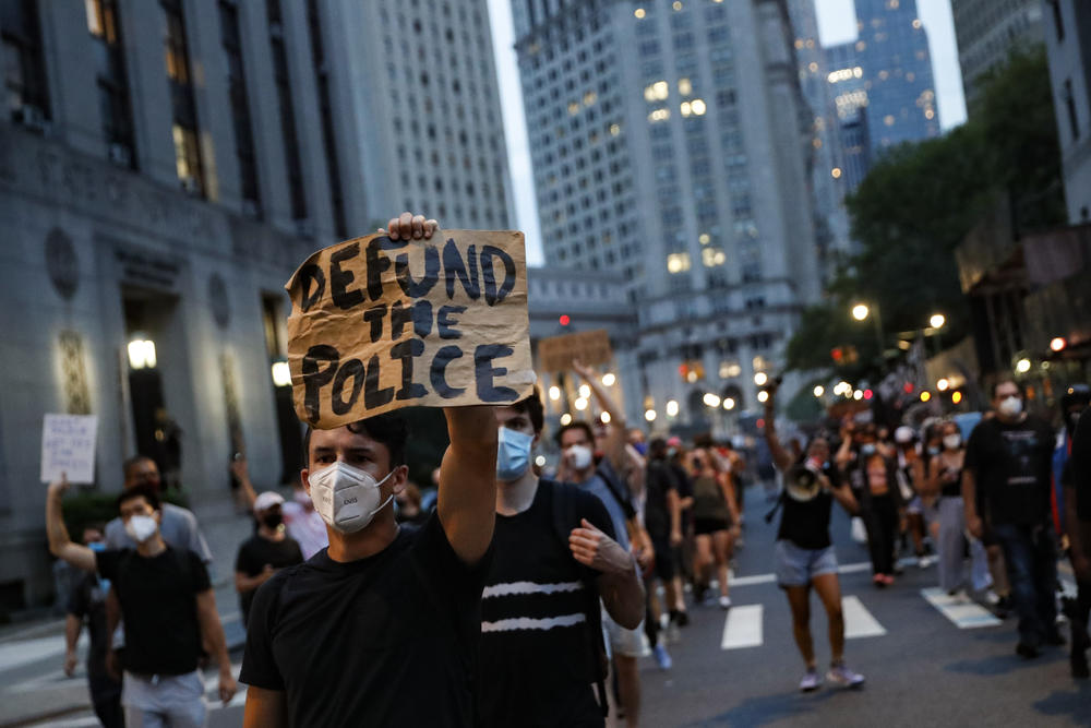 A man at a protests holds a cardboard sign that reads, "Defund the Police."