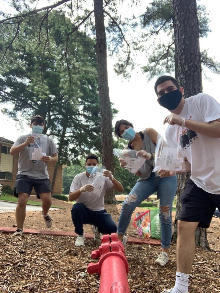 Four college students wearing masks hold plastic bags with supplies and information on COVID-19. Behind them, a pine tree. 