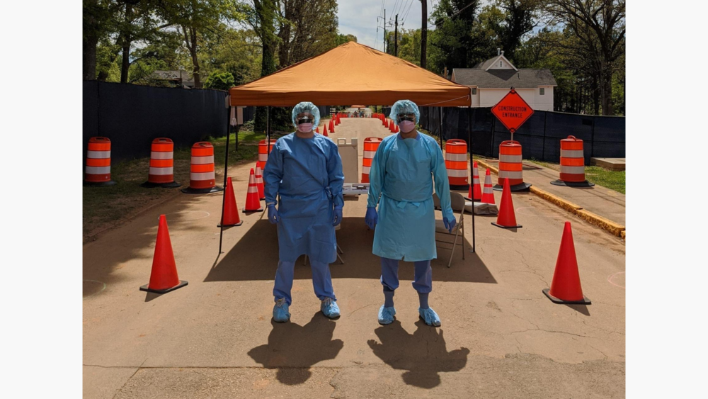 Two people are in the foreground with blue hairnets, blue medical scrubs, and a white mask looking at the camera. On the left and right, construction cones fade into the background. An orange medical aid tent station is behind them.