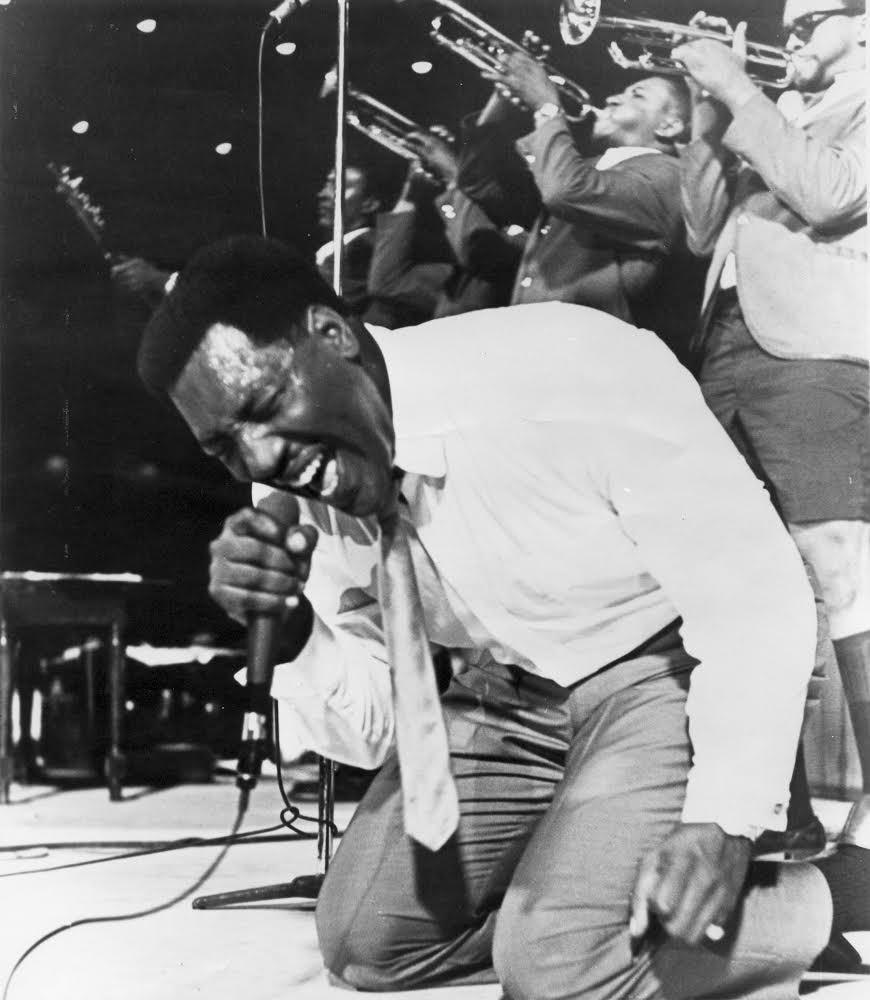 Otis Redding on his knees, singing into a microphone. Behind him, three men play trumpets.