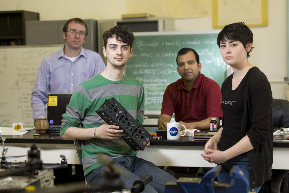 Post Doc research scientist David Cotten of the Center for Geospatial Research, Senior computer science and astrophysics major Caleb Adams, Professor of Geography Deepak Mishra, and senior mechanical engineering major Megan Le Corre are working together along with a team of students and professors in a collaborative effort to design, build, and deploy (with the help of NASA) a cube satellite. Adams is holding a 3D printer model of the CubeSat.