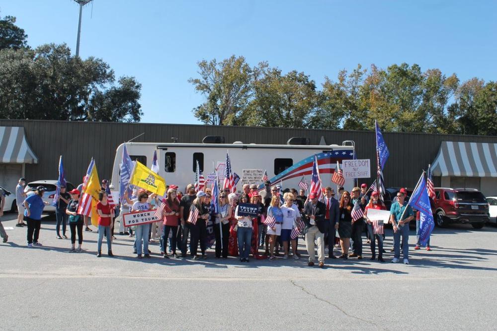 A caravan of trucks, all bearing out-of-state license plates and oversized Trump flags, was parked near the crowd, which gathered in front of an RV that had “TRUMP GIRLS” emblazoned on its front windshield.