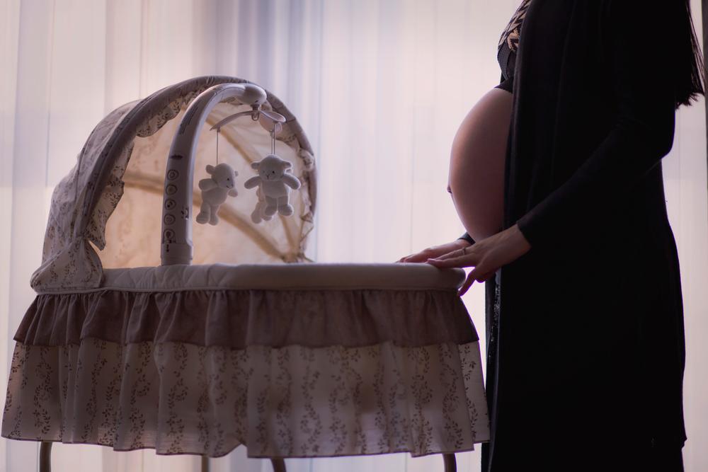 A pregnant woman stands next to a crib.
