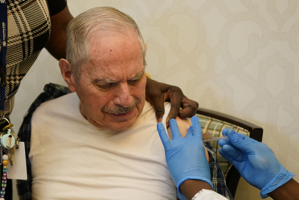 An older man gets a shot into his left arm while sitting on an armchair.