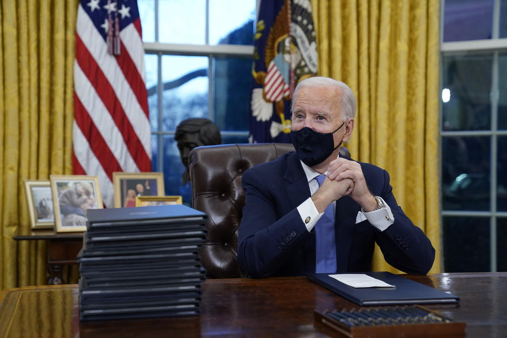 President Joe Biden sits in the oval office next to a stack of executive orders.