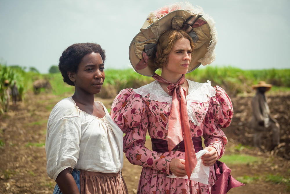 Two women, one in elaborate 19th century dress and one in a simple blouse and skirt, stand in a sugarcane field.