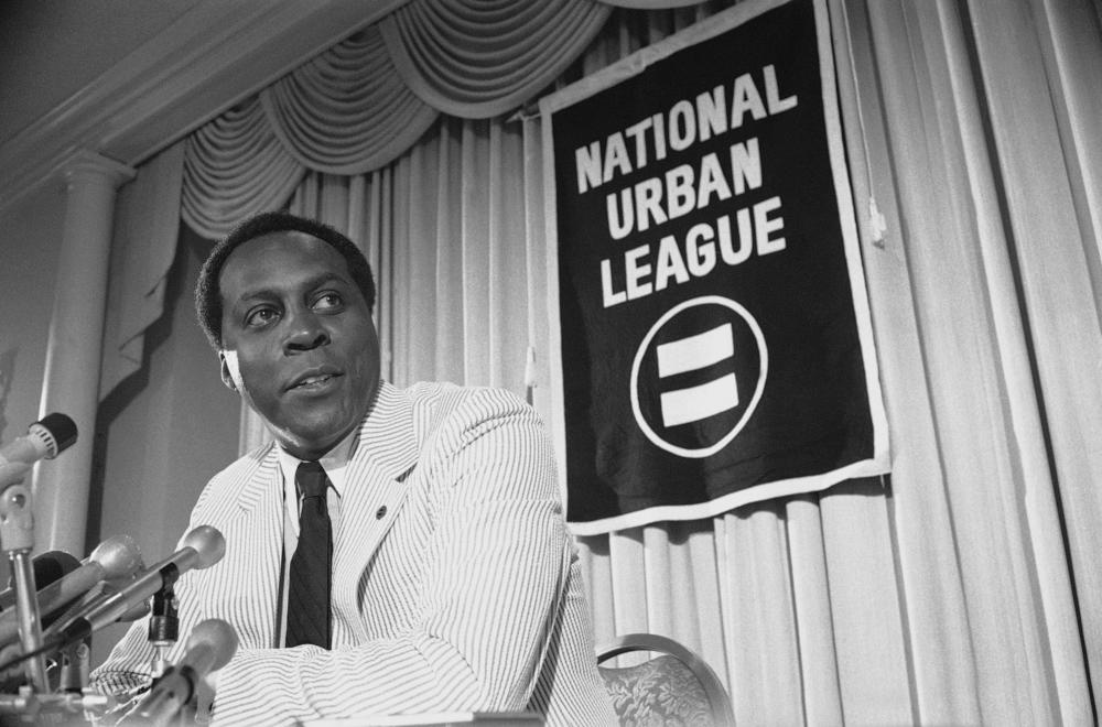 A black and white photo of Vernon Jordan sitting at some microphones during a press conference with the National Urban League.
