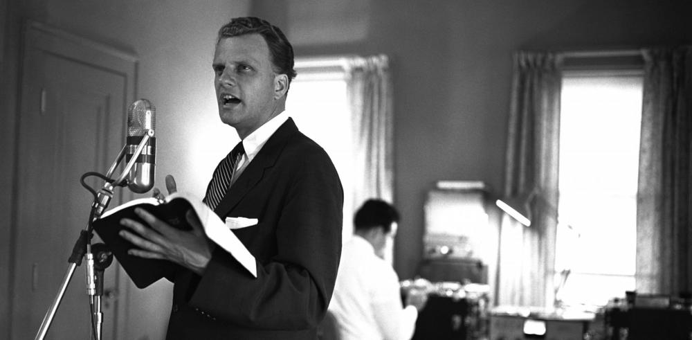 Billy Graham preaches at a microphone, holding a Bible. 1959.