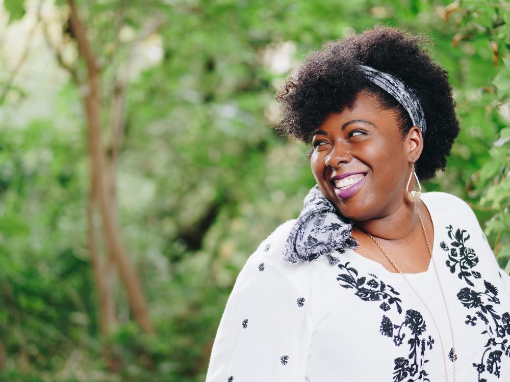 A Black woman looks over her shoulder outdoors with trees behind her.