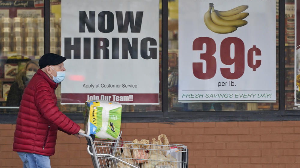 A man with a face mask covering only his mouth pushes a grocery cart outside a store window with a now hiring sign.