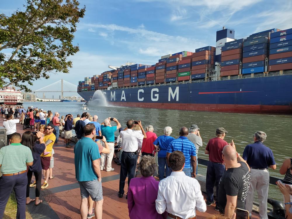 an enormous container ship passes a crowd of people watching from the shore