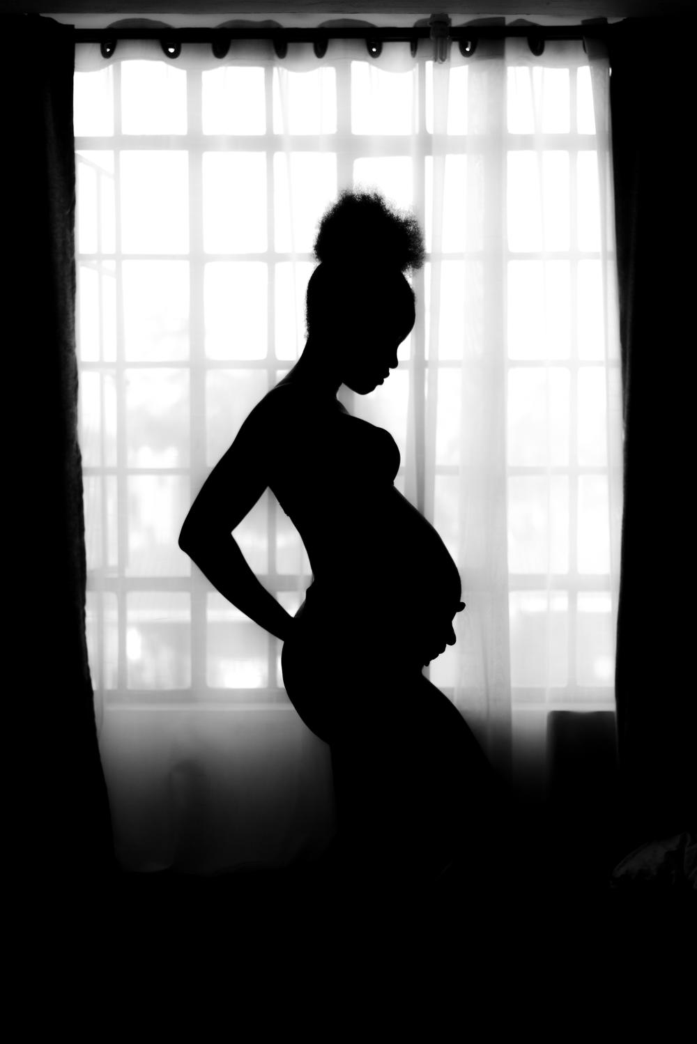 A pregnant woman stands holding her belly in front of a window.