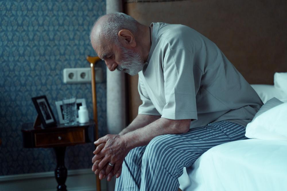 A man sits on the edge of a bed, looking down.
