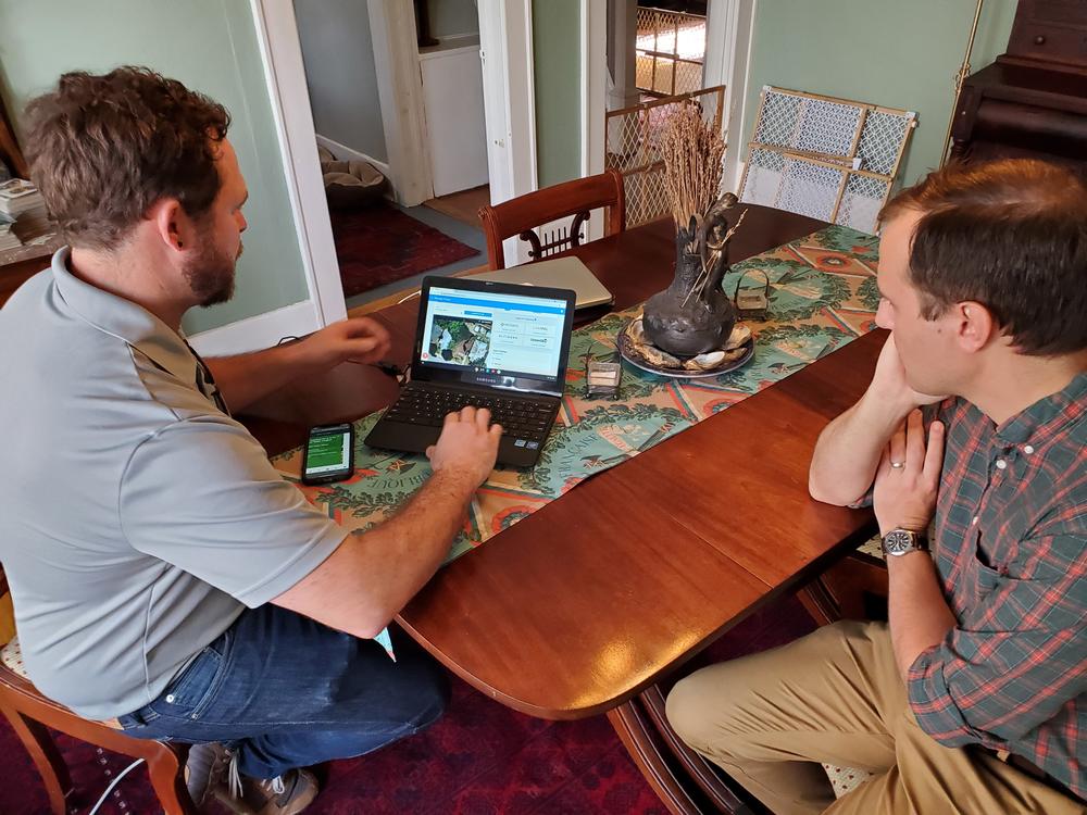 two men sit at a dining table, looking at a laptop