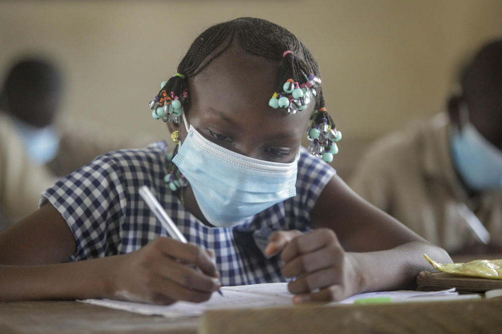 A girl in a mask writes on paper at a school desk. 