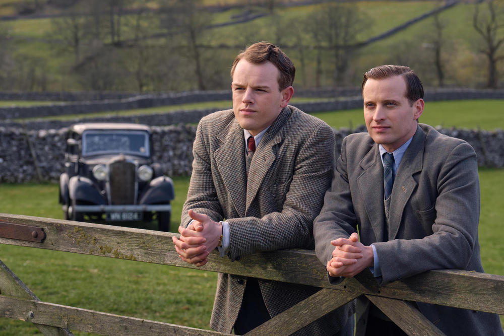 Two men lean against a wooden fence.