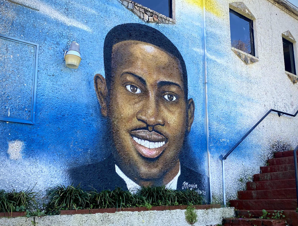 A mural of Ahmaud Arbery, on the side of a building in Brunswick. Arbery is smiling, and wearing a black tuxedo and white collared shirt.