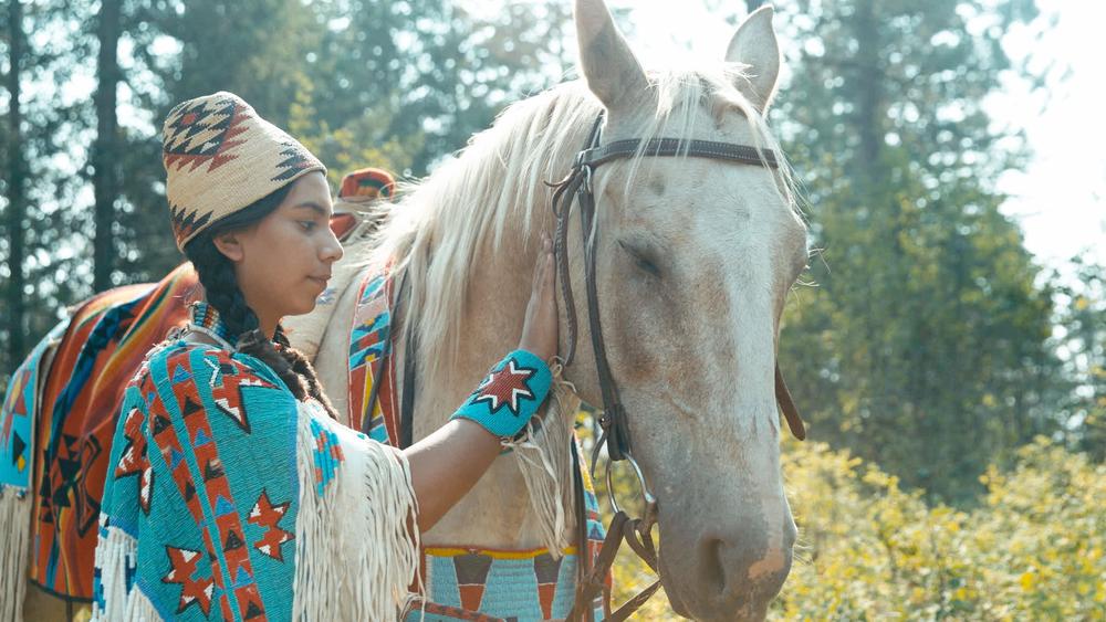 Close up of Nez Perce girl dressed in regalia, petting her Appaloosa horse in the forest. Nez Perce Tribal Land, Idaho.