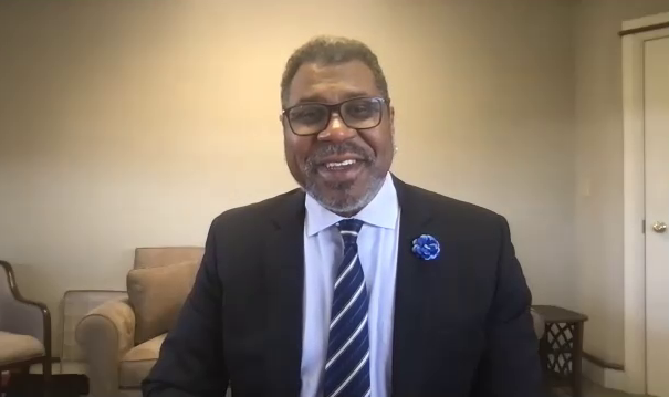 A screenshot from a Zoom interview with Gene Dobbs Bradford, the new executive director of the Savannah Music Festival. He is sitting in his office, wearing a black suit and smiling at the camera.