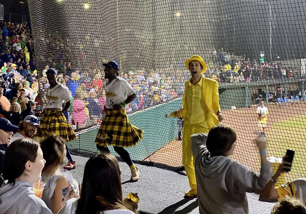 Wearing his signature yellow tuxedo, Savannah Bananas owner Jesse Cole dances with players and fans in between innings at Grayson Stadium. Cole and two players are dancing on top of the home dugout. The players are wearing kilts with a yellow-and-black plaid pattern, in addition to their white home uniforms.