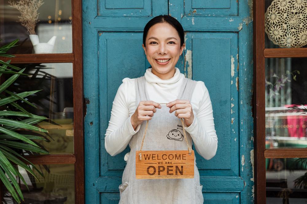 A female cafe owner holds a sign that says WELCOME, WE ARE OPEN