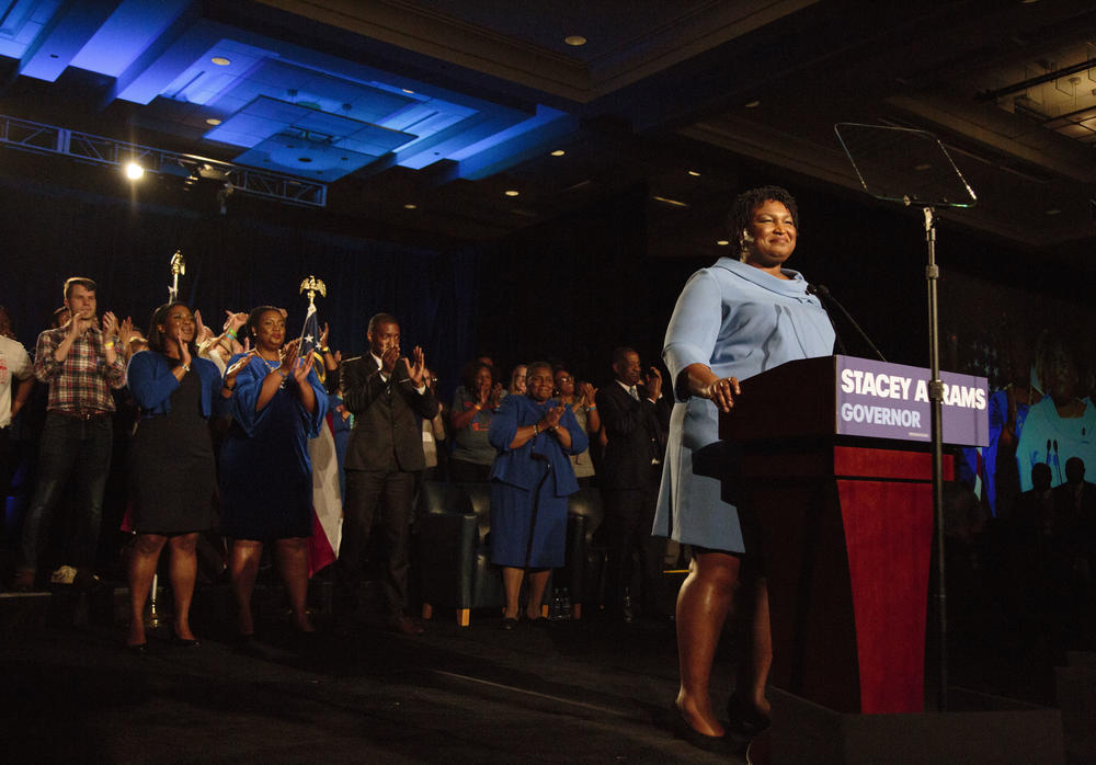 Stacey Abrams stands on stage with supporters clapping behind here.