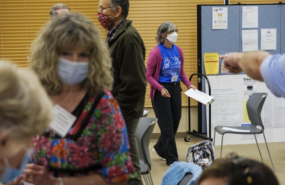 A scene from an election polling site shows people waiting in line and a poll watcher carrying a clipboard.