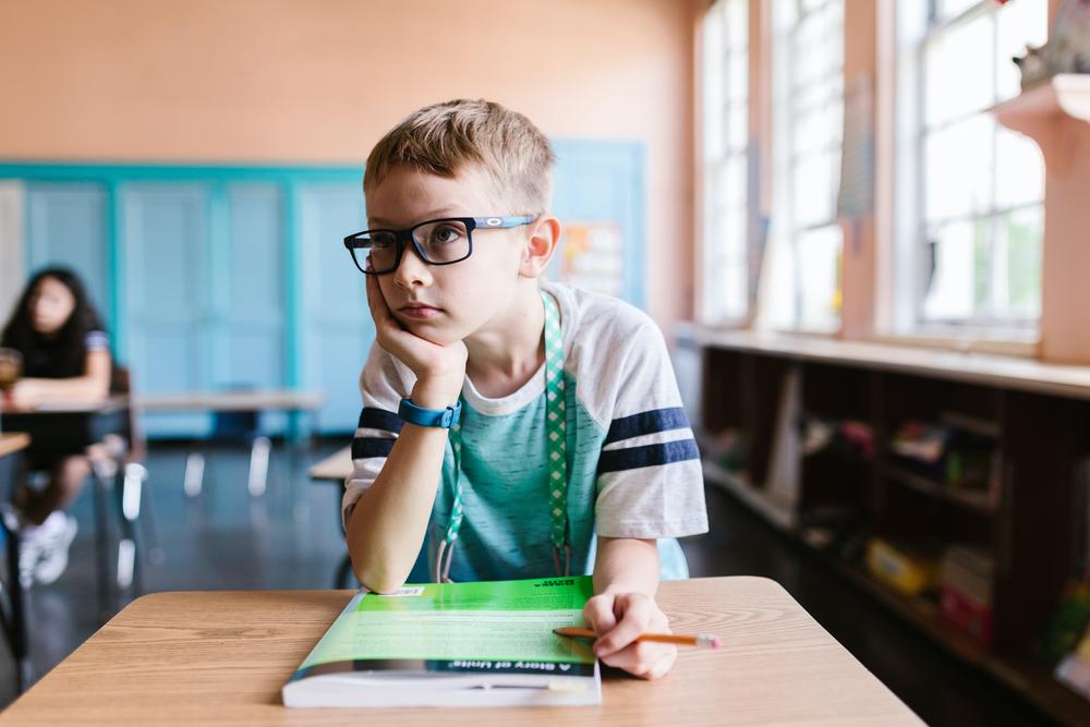 A boy with glasses sits in class with his hand on his cheek